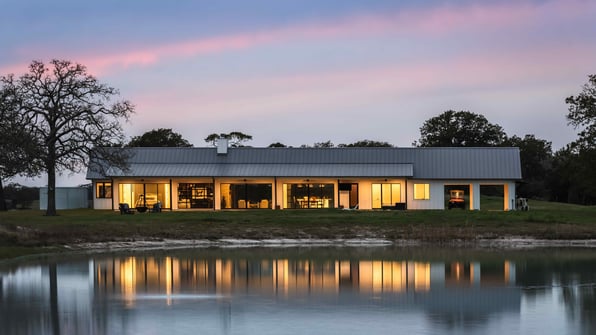 Modern Texas Hill Country home designed by J. Bryant Boyd, with a standing-seam metal roof and glass walls glowing at dusk beside a pond