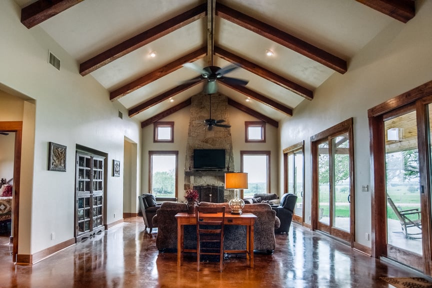 Vaulted living room in a Hill Country custom home by J. Bryant Boyd, with exposed timber beams, a stone fireplace, and stained concrete floors facing pasture views
