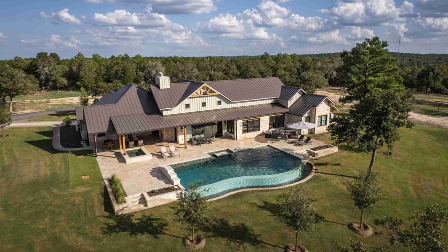 Aerial view of a sprawling custom home with metal roofing, stone exterior, and a uniquely-shaped pool surrounded by open fields. In Texas Hill Country | J Bryant Boyd